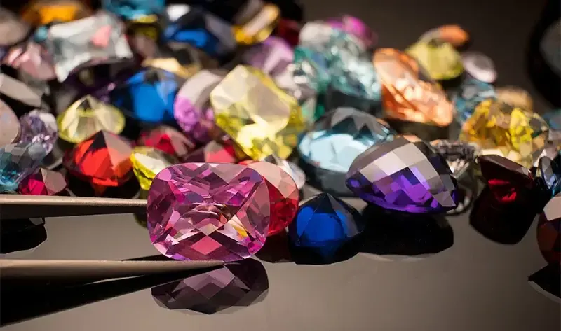 Assorted faceted colored gemstones with a stone held in tweezers