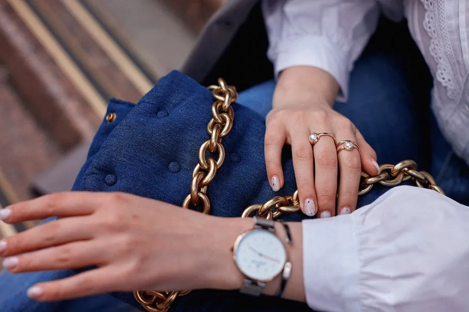 Hand wearing two rings with pearl-like stones beside a blue handbag